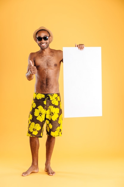 Retrato de un hombre afro feliz sosteniendo un tablero en blanco y mostrando un signo bien aislado en una pared naranja