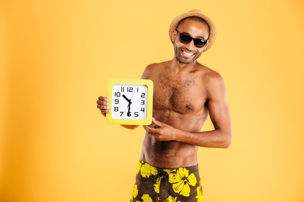 Retrato de un hombre afro feliz en traje de baño con reloj de pared aislado en una pared naranja