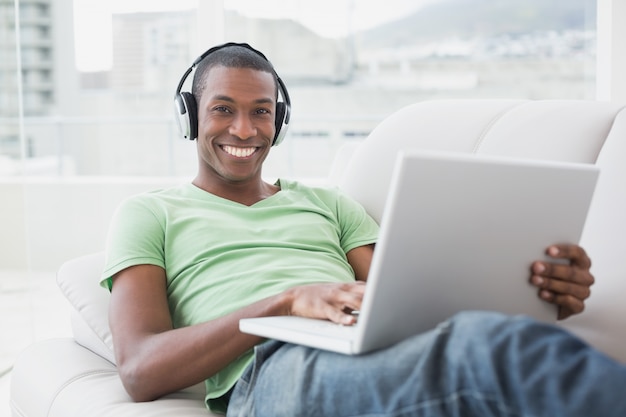 Retrato de hombre afro sonriente con auriculares usando la computadora portátil en el sofá