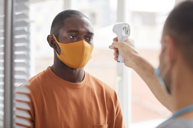 Retrato de hombre afroamericano adulto con máscara obteniendo control de temperatura mientras espera en línea en la clínica