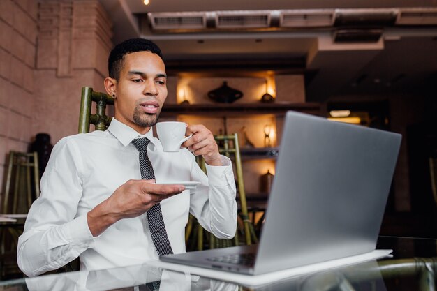 Retrato de un hombre afroamericano beber café y trabajar en un portátil, con una camisa blanca, interior