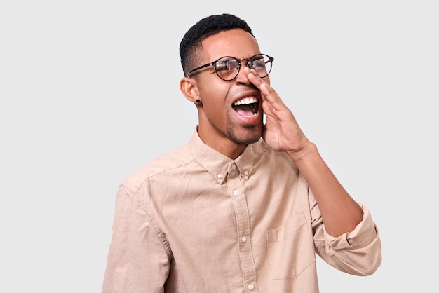 Retrato de un hombre afroamericano con una camisa beige de gafas redondas gritando con la boca abierta Un hombre furioso de piel oscura gritando en voz alta posando en la pared blanca del estudio
