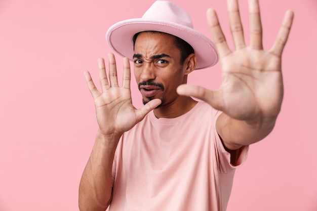 Retrato de hombre afroamericano confundido con sombrero mostrando gesto de parada aislado en rosa