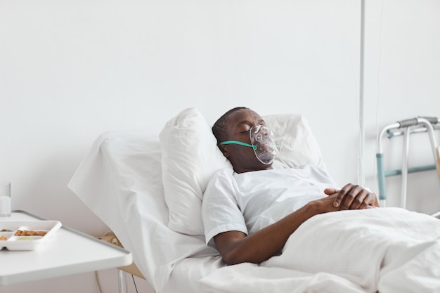 Foto retrato de hombre afroamericano durmiendo en la cama de un hospital con máscara de oxígeno en la habitación blanca, espacio de copia