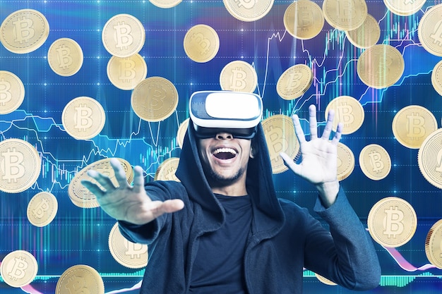 Retrato de un hombre afroamericano emocionado con una capucha negra y gafas vr. Hay bitcoins y gráficos en el fondo.