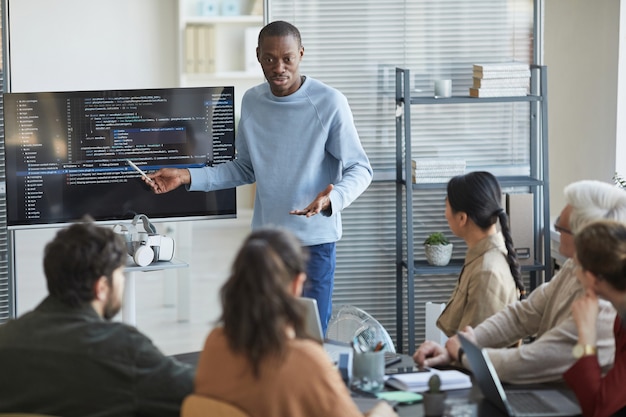 Retrato de hombre afroamericano emocional dando presentación en la oficina al equipo de TI y apuntando al código en la pantalla, espacio de copia