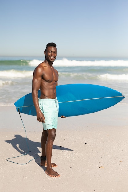 Un retrato de un hombre afroamericano feliz y atractivo que disfruta del tiempo libre en la playa en un día soleado, sonriendo, divirtiéndose, de pie con su tabla de surf, el sol brillando sobre él.
