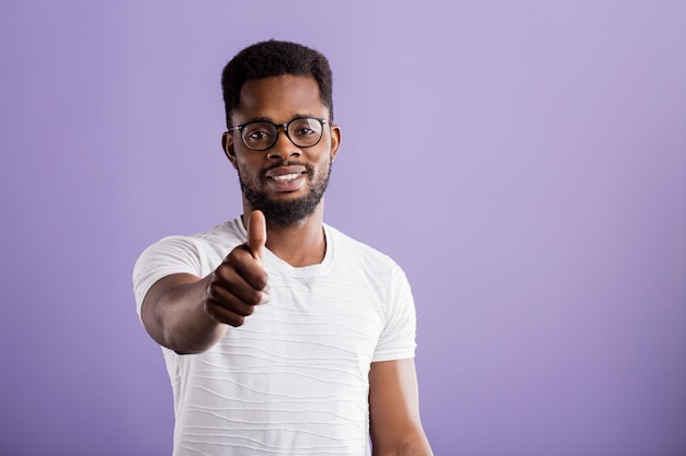 Retrato de hombre afroamericano guapo satisfecho con barba en gafas, camiseta blanca casual que muestra el pulgar hacia arriba sonriendo con expresión feliz sobre fondo lila. De cerca, copie el espacio.