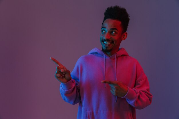 Retrato del hombre afroamericano sonriente en la colorida sudadera con capucha mirando y señalando con el dedo a un lado aislado sobre la pared violeta