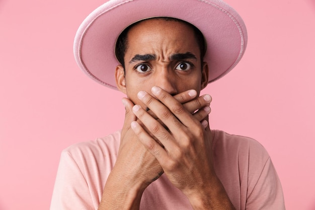 Retrato de hombre afroamericano sorprendido con sombrero coning su boca aislada en rosa