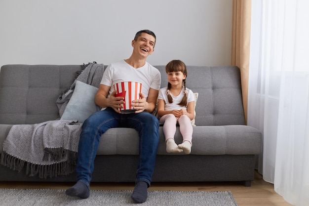 Retrato de un hombre agradable, encantador, positivo y alegre que usa pantalones vaqueros casuales de camiseta blanca sentado en un sofá divirtiéndose viendo videos divertidos con su hija disfrutando de pasar el tiempo libre