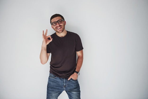 Retrato de un hombre alegre en camiseta y anteojos y mostrando el signo de ok mientras mira a la cámara sobre fondo blanco.