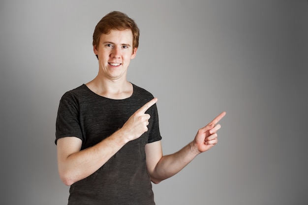 Retrato de hombre alegre joven pelirrojas sonriendo apuntando con el dedo hacia arriba. Sobre pared gris. Copie el espacio.