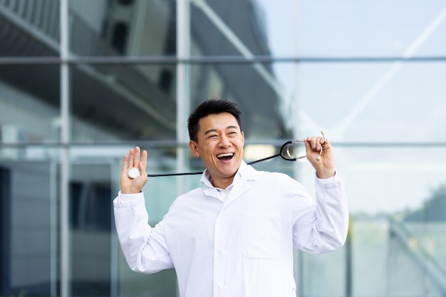 Retrato de un hombre alegre médico asiático bailando feliz con los resultados del trabajo realizado en el fondo de una clínica moderna en el exterior