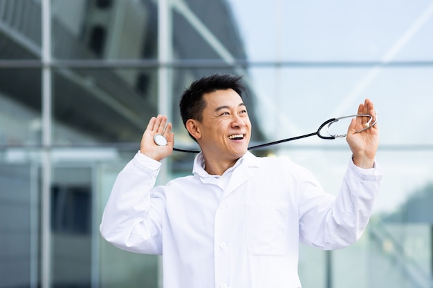 Retrato de un hombre alegre médico asiático bailando feliz con los resultados del trabajo realizado en el fondo de una clínica moderna en el exterior