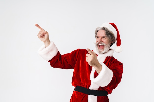 Retrato de hombre alegre de pelo gris de Santa Claus en traje rojo apuntando con el dedo a un lado en copyspace aislado en blanco