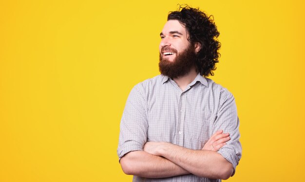 Retrato de hombre alegre rizado y barbudo de pie sobre fondo amarillo con los brazos cruzados