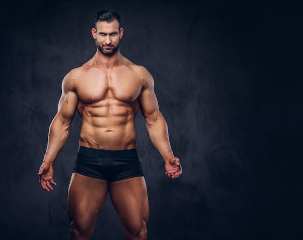 Retrato de un hombre alto y sin camisa con un cuerpo musculoso con un elegante corte de pelo y barba, en ropa interior, posando en un estudio. Aislado en un fondo oscuro.