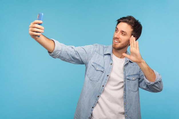 Retrato de un hombre apuesto y amigable con camisa de mezclilla hablando por videollamada y saludando con un gesto de hola teniendo una conversación en línea en el teléfono móvil tomando una foto de estudio selfie aislada en el fondo azul