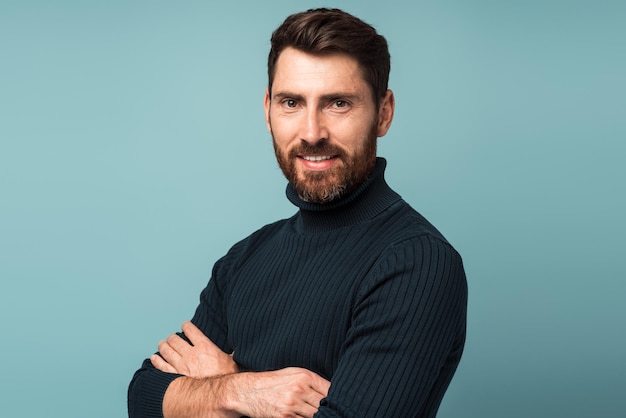 Retrato de un hombre apuesto y barbudo con un estilo informal mirando a la cámara y sonriendo mientras posa con los brazos cruzados Foto de estudio sobre fondo azul