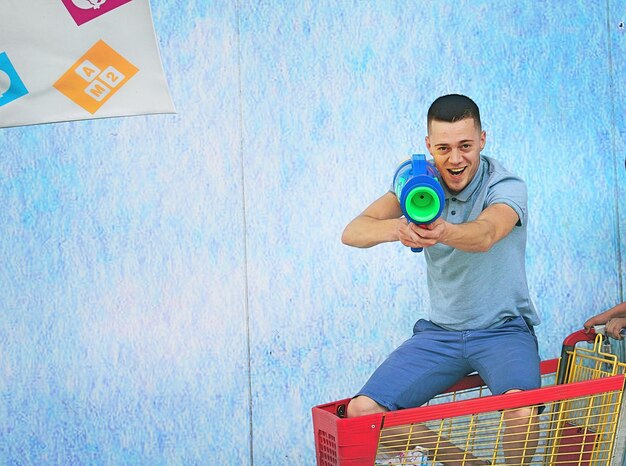 Foto retrato de un hombre apuntando con una pistola de agua en un carrito de compras junto a la pared