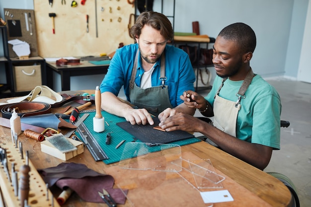 Retrato de hombre artesano enseñando joven aprendiz en silla de ruedas en el taller de peleteros copia ...