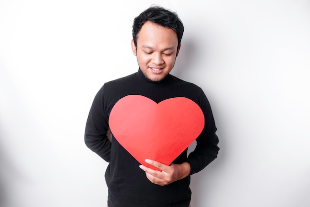 Un retrato de un hombre asiático feliz con una camisa negra sosteniendo un papel rojo en forma de corazón aislado por un fondo blanco