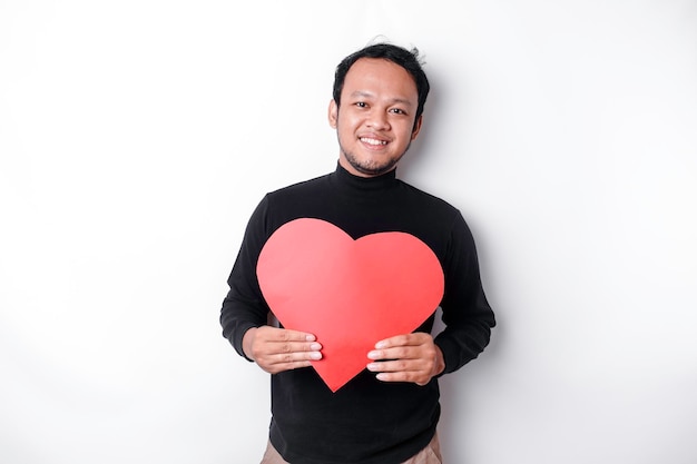 Un retrato de un hombre asiático feliz con una camisa negra sosteniendo un papel rojo en forma de corazón aislado por un fondo blanco