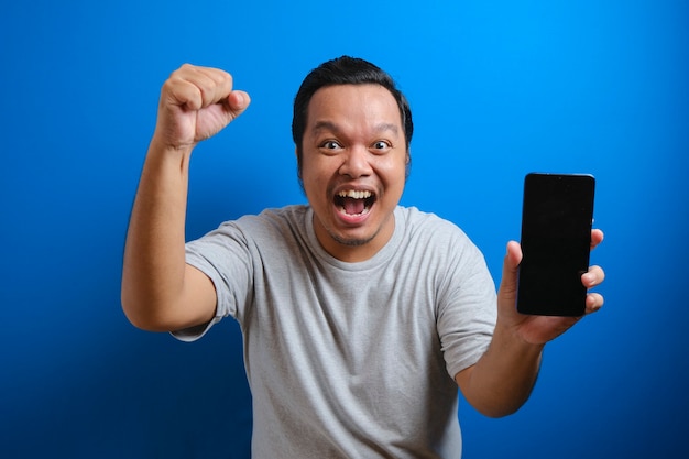 Retrato de hombre asiático gordo sorprendido en camiseta gris apuntando al teléfono celular y mirando a la cámara con asombro. El hombre muestra un gesto ganador levantando la mano. Foto de estudio aislado sobre fondo azul.