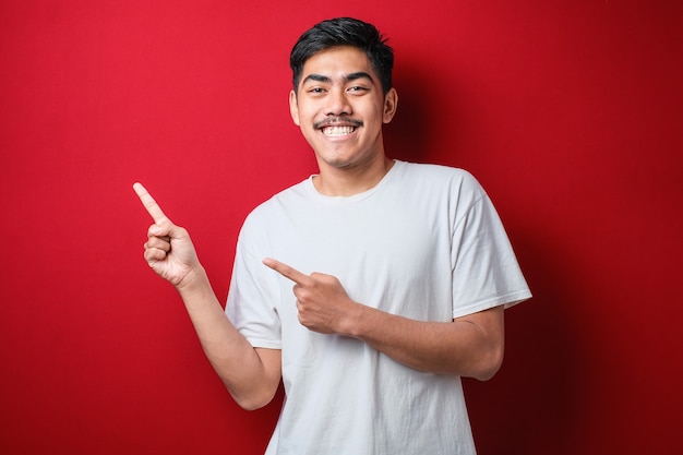 Retrato de hombre asiático joven divertido en camiseta blanca sonriendo y apuntando a presentar algo de su lado, contra un fondo rojo con espacio de copia