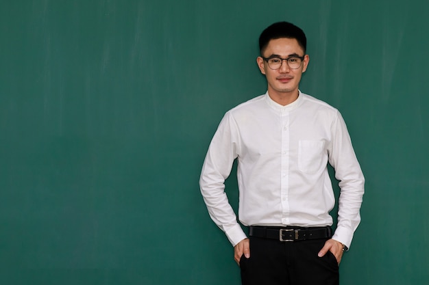 Retrato de hombre asiático joven y guapo con anteojos y ropa casual de negocios, camisa blanca y pantalón negro, pose de pie con confianza en sí mismo con fondo verde y espacio de copia.