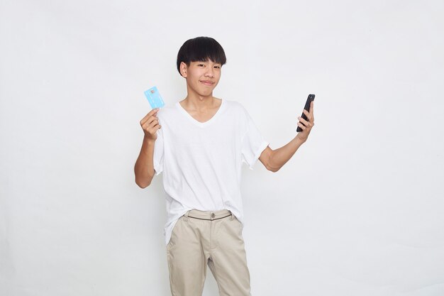 Retrato de un hombre asiático joven lindo que sostiene el teléfono móvil y que muestra la superficie blanca aislada de la tarjeta de crédito