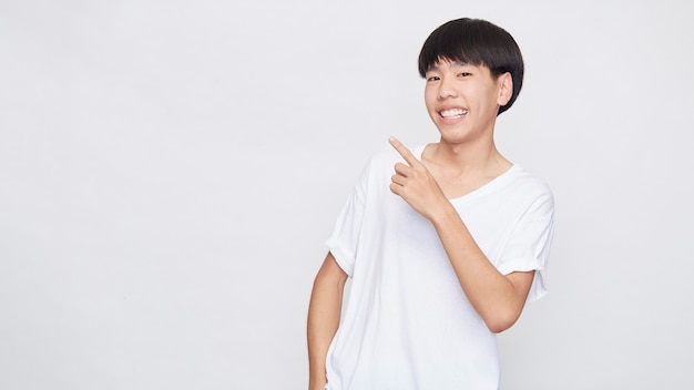 Retrato de un hombre asiático sonriente feliz vestido con camiseta blanca casual apuntando con la mano al espacio vacío al lado sobre fondo blanco.