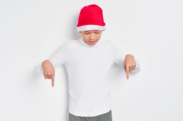 Retrato de un hombre asiático sonriente con sombrero de navidad señalando con el dedo hacia abajo invitando a la gente a seguir aislado en el fondo blanco