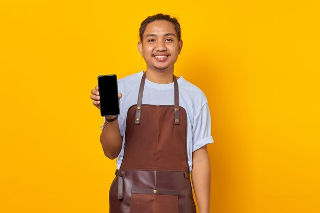 Retrato del hombre asiático sonriente vistiendo delantal mostrando la pantalla en blanco del smartphone sobre fondo amarillo