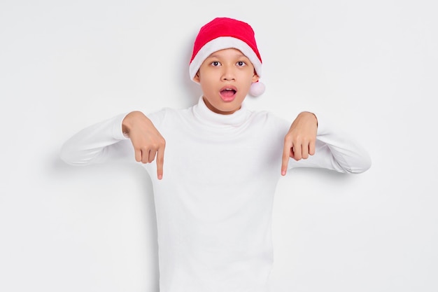 Retrato de un hombre asiático sorprendido con sombrero de Navidad señalando con el dedo hacia abajo invitando a la gente a seguir aislado de fondo blanco