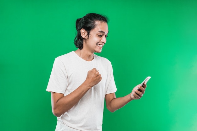 Retrato de hombre asiático sosteniendo y mirar telefono con expresión de felicidad mientras levanta una mano y hace un puño, aislado sobre fondo verde