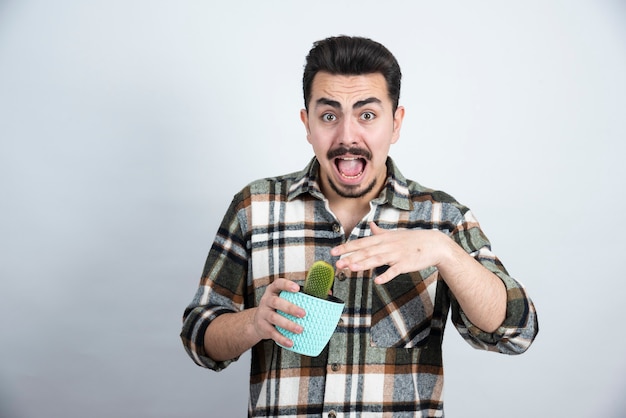 Retrato de hombre asustado manteniendo pequeño cactus en maceta azul sobre pared blanca.