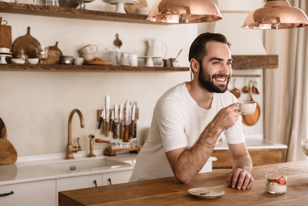 Retrato de hombre atractivo morena comiendo postre con cucharadita mientras desayuna en la elegante cocina de casa