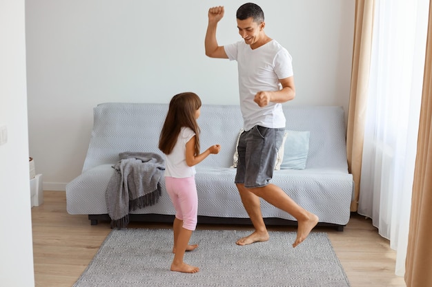 Retrato de hombre atractivo positivo con camiseta blanca y jeans cortos bailando con su hija en el interior cerca del sofá en la sala de estar, celebrando las vacaciones y divirtiéndose juntos.
