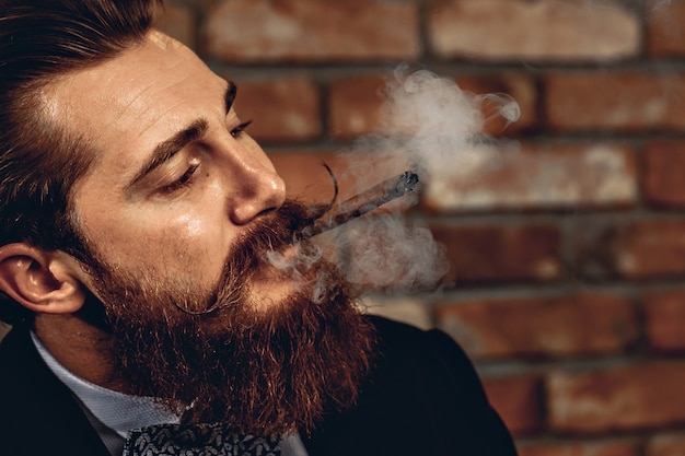 Retrato de un hombre atractivo sharming con un bigote de húsar y barba fumando un puro marrón y el humo vuela por la habitación. Concepto de adicción