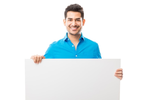 Retrato de hombre atractivo sonriendo mientras sostiene un cartel en blanco contra un fondo completamente blanco