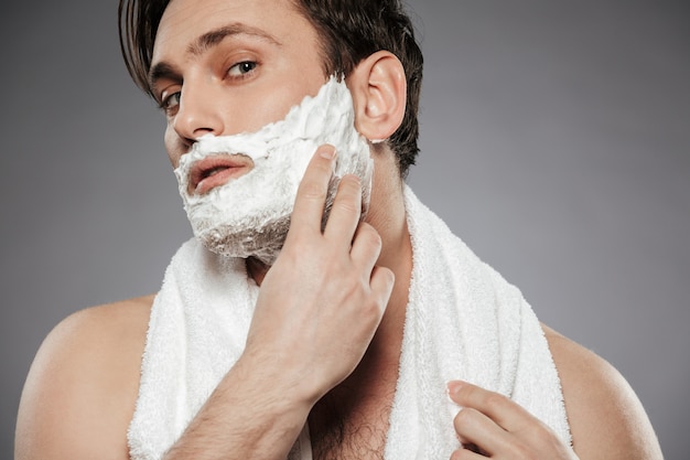 Retrato de hombre atractivo con una toalla en el cuello poniendo espuma de afeitar en la cara, mientras que el cuidado de la piel por la mañana aislado sobre la pared gris