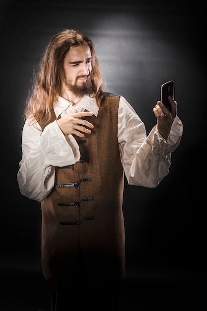 Retrato de un hombre con barba y cabello largo vistiendo un traje de pirata medieval en un espacio negro, un pirata sosteniendo un teléfono móvil