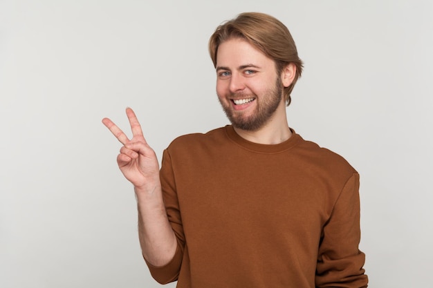 Retrato de hombre con barba con sudadera haciendo gesto de victoria y sonriendo a la cámara mostrando el signo de paz v con dedos dobles Estudio interior aislado sobre fondo gris