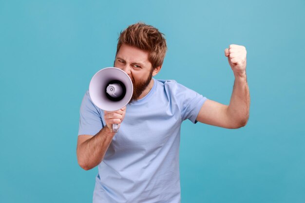 Retrato de un hombre barbudo agresivo sosteniendo un megáfono cerca de la boca, hablando en voz alta, gritando, haciendo anuncios con el brazo levantado, protestando. Disparo de estudio interior aislado sobre fondo azul.