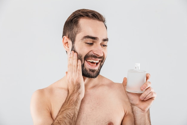 Retrato de un hombre barbudo sin camisa alegre aplicar loción para después del afeitado aislado sobre blanco