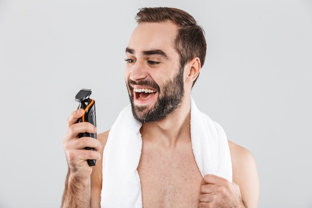 Retrato de un hombre barbudo sin camisa alegre que muestra la maquinilla de afeitar eléctrica aislada sobre blanco