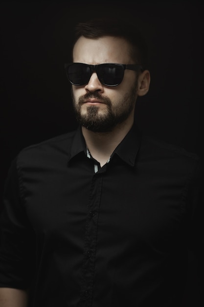 Retrato de un hombre barbudo con una camisa negra y gafas de sol con una mirada intensa de pie en el interior