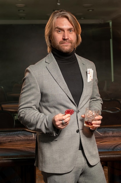 Retrato de un hombre barbudo con experiencia y confiado listo para jugar al póquer en el casino, parado cerca de la mesa de juego con un vaso de bebida y fichas de apuestas en las manos